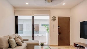 A living room with a sofa, a TV, a door, and a view into another room. - Oceanview Rooftop Pool, Gym & 1 Block to 5th Ave (Playa del Carmen)