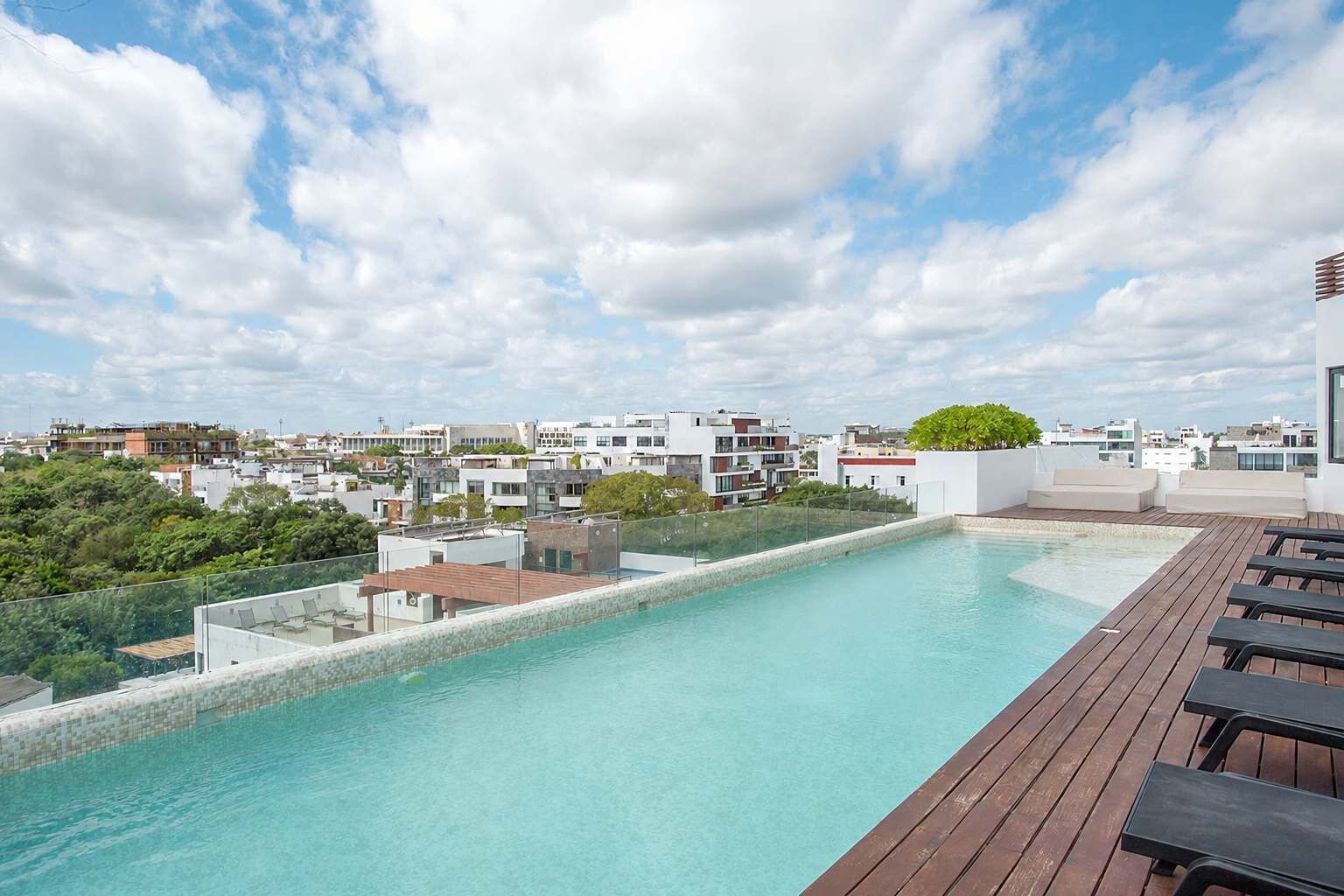 A rooftop pool with a wooden deck and lounge chairs, surrounded by modern buildings and greenery.
