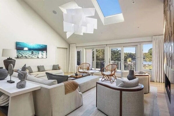 A modern living room with a high ceiling, large windows, and a skylight. There is a sofa, chairs, and a coffee table.