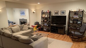 A living room with a sectional sofa, a TV, and a bookshelf. - Resort like Backyard-Pool, Putting green, Outside Kitchen, Fire Pit, Bar, Lounge (Hampton Bays)