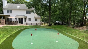 A house with a front yard featuring a green artificial turf putting green with three red golf holes. - Resort like Backyard-Pool, Putting green, Outside Kitchen, Fire Pit, Bar, Lounge (Hampton Bays)