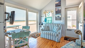 Une pièce avec un plancher en bois, un canapé à rayures, une table à manger avec des chaises, un téléviseur et une vue sur la plage grâce à de grandes fenêtres.