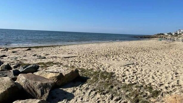 A beach with a rocky shoreline, clear blue sky, and buildings in the background. - Newly renovated . Near sandy association beach RPMS090 (Dennis Port)