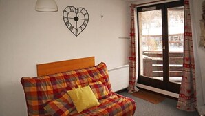 Huoneessa on ruutukuviollinen sohva, keltainen tyyny, sydämenmuotoinen kello ja ikkunasta avautuva näkymä rakennukseen, ja ikkunassa on kuviolliset verhot.