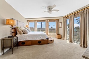 A bedroom with a large bed, a ceiling fan, a nightstand, and a view of the outside through large windows.