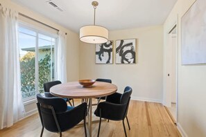 A dining area with a round table, four chairs, a hanging light fixture, and two abstract paintings on the wall.