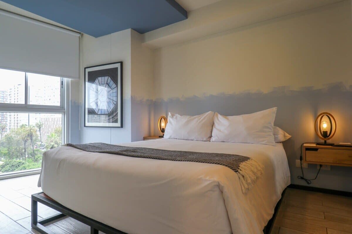 A modern hotel room with a large bed, a framed artwork on the wall, and a view of the city through the window.