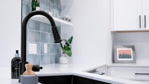 Une cuisine moderne avec un robinet noir, un plan de travail blanc et un évier noir. On peut voir une bouteille de savon Bayla et une petite plante sur le plan de travail.