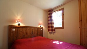 Une chambre à coucher avec une tête de lit en bois, deux lampes de chevet, une fenêtre avec des rideaux à carreaux et une couvre-lit rouge.