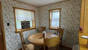 A dining area with a round wooden table and four chairs, two windows with curtains, and a wall with a floral pattern.