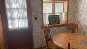 A room with a wooden door, a window with a sign, a round wooden table, and four wooden chairs.