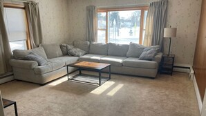A living room with a beige carpet, a grey sectional sofa, a small coffee table, and a lamp on a side table.