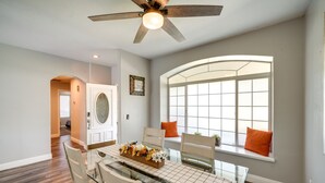 Comedor con mesa de vidrio, sillas, ventilador de techo y ventana con cojines naranja.