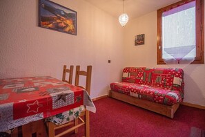Ein Zimmer mit einem Holztisch und einem roten Sofa mit Steppbezug. An der Wand hängt ein Gemälde und das Fenster bietet einen Blick auf eine Landschaft.