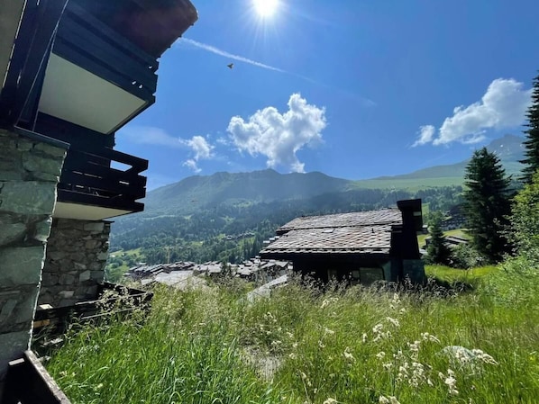A stone building with a sloped roof, surrounded by lush green fields and mountains in the background.