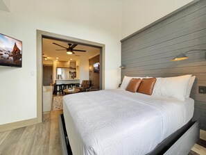 Une chambre d’hôtel avec un grand lit, un ventilateur de plafond, une tête de lit en bois et un coin repas visible à travers une porte ouverte.