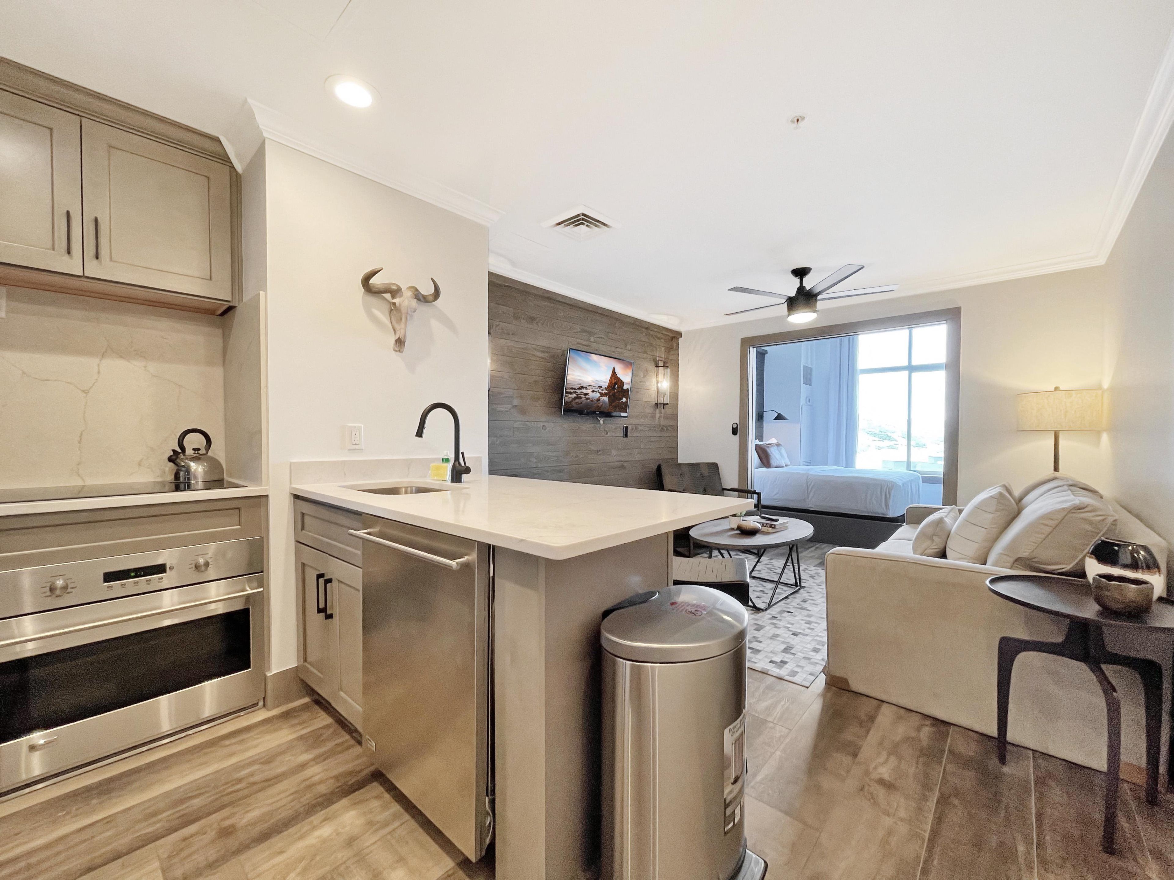 A modern kitchen with stainless steel appliances, a dining area with a round table, and a living space with a sofa and a TV mounted on the wall.