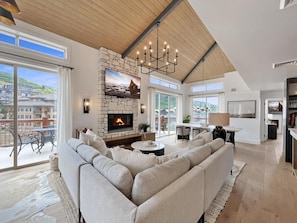 A spacious living room with a stone fireplace, a large flat-screen TV, and a high ceiling with exposed beams.