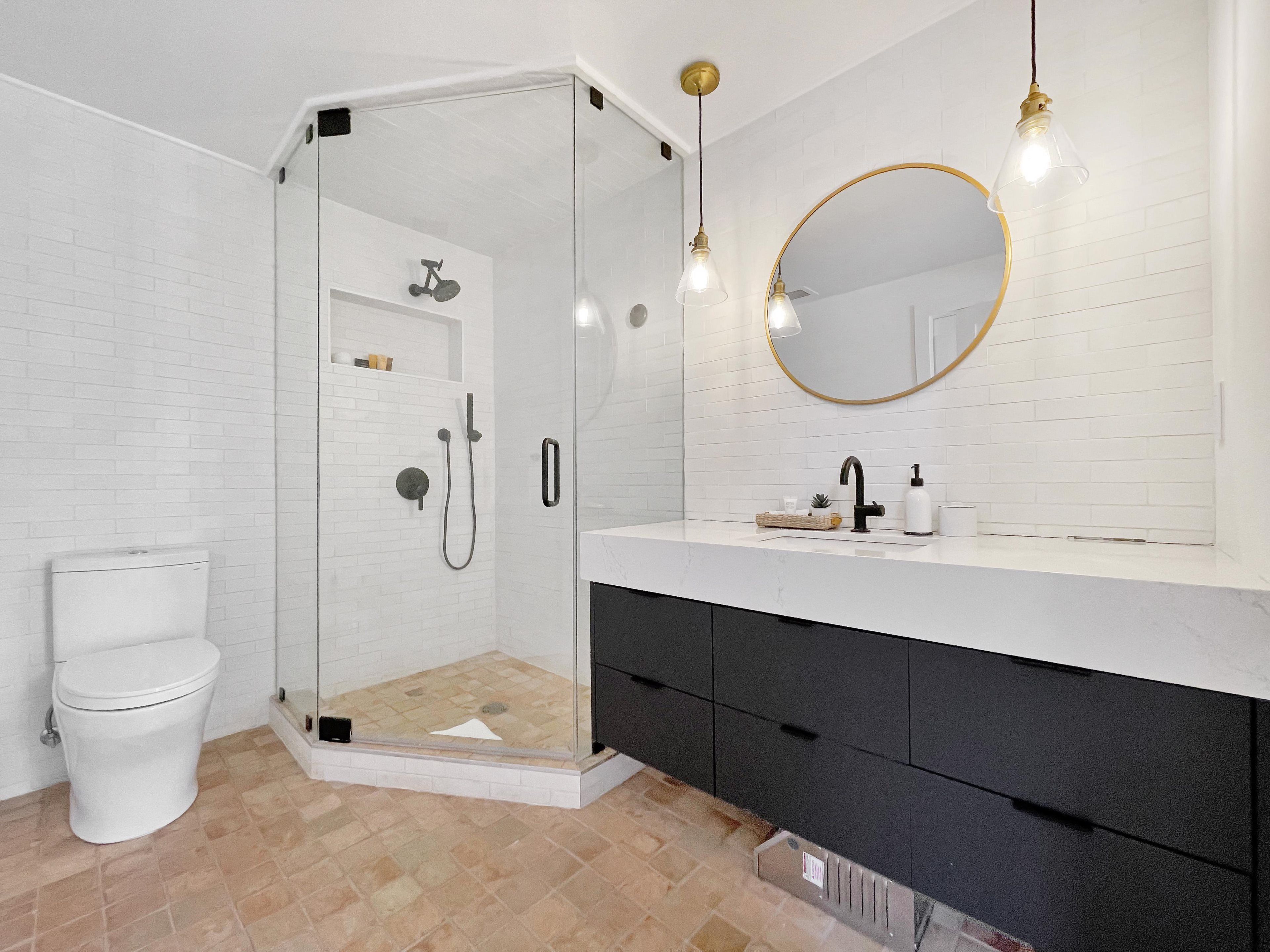 A modern bathroom with a glass-enclosed shower, a white toilet, a marble countertop, a black vanity, and a round mirror with a gold frame.