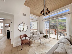 Un vaste espace de vie comprenant un canapé, un fauteuil et une table basse, un grand miroir rond accroché au mur, et un balcon offrant une vue sur les immeubles.