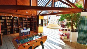 A spacious indoor area with wooden bookshelves, a foosball table, and a view of a courtyard with a fountain. - La Petite Fecht (Munster)