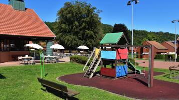 A playground with a slide and climbing area, surrounded by a grassy area with benches and tables.