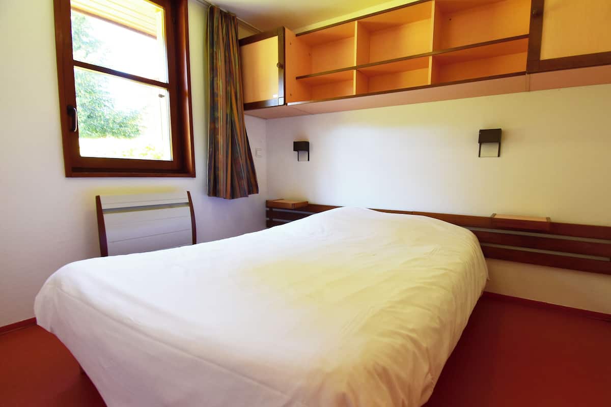 A hotel room with a bed, a chair, a window, and built-in shelves.