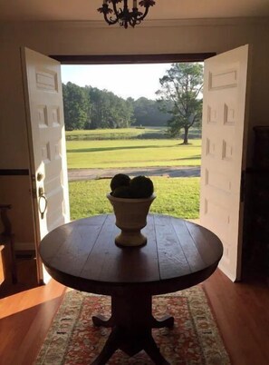 Eine offene Tür, die auf einen Blick auf einen Golfplatz freigibt, ein runder Holztisch mit einem Topfpflanzen und ein gemustertes Teppich auf dem Boden.