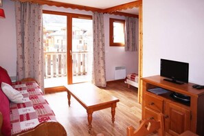 Ein Zimmer mit Holzboden, einer Couch, einem Fernseher auf einem Holzschrank und einem Fenster mit Vorhängen.