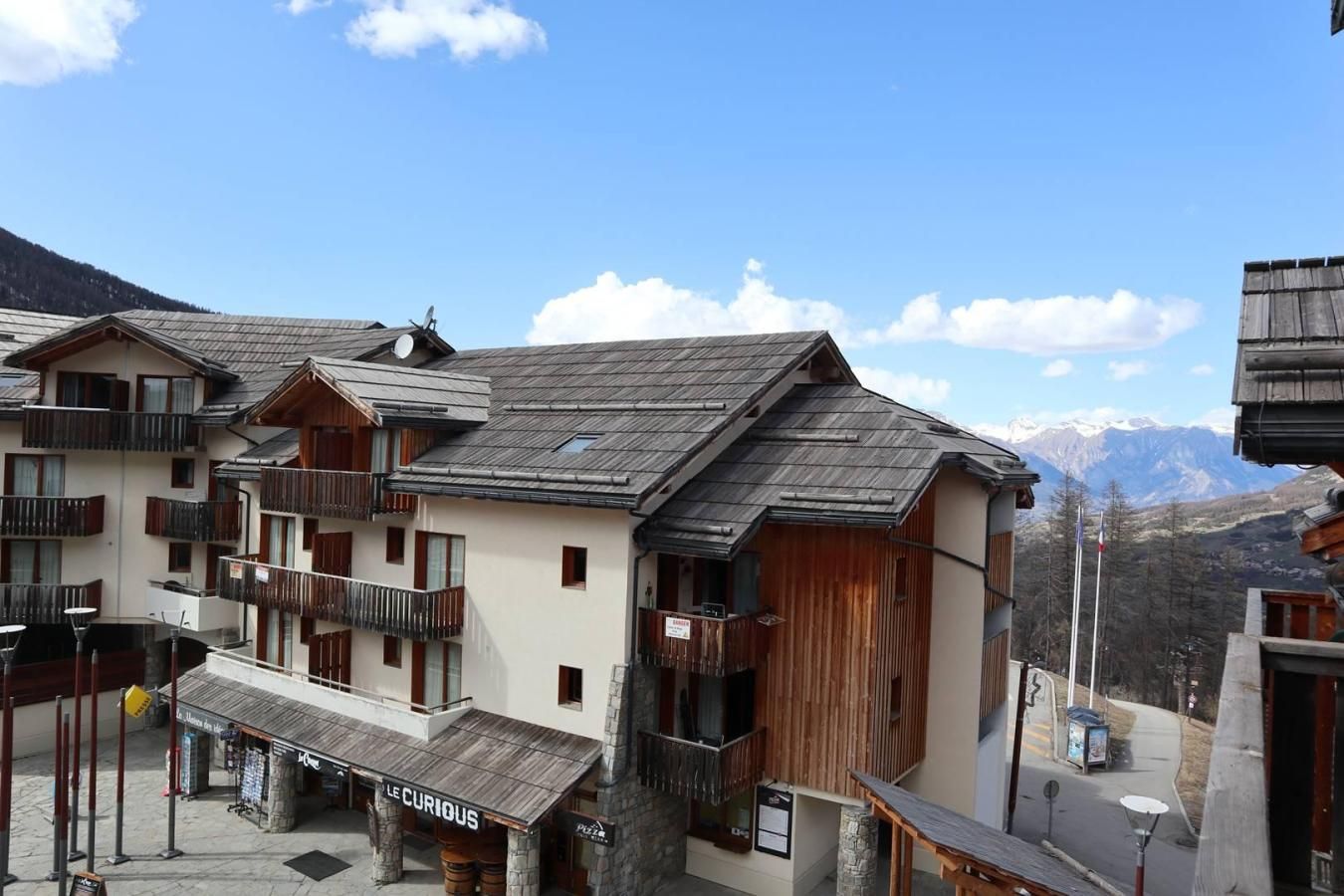 A mountain resort with chalet-style buildings, balconies, and a restaurant named 'Le Curieux'.