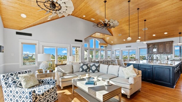 A spacious living room with a large sofa, a coffee table, and a dining area with a view of the ocean. The kitchen is visible in the background with dark cabinetry and a large island. - Luxury Semi-Oceanfront Home w/ Pool, Elevator, Theater Room, and Game Room (Hatteras)
