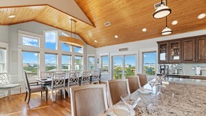 A spacious kitchen with a large island, wooden ceiling, and large windows offering a view of the outdoors. - Luxury Semi-Oceanfront Home w/ Pool, Elevator, Theater Room, and Game Room (Hatteras)