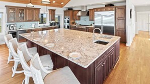 A spacious kitchen with a large island, wooden cabinets, stainless steel appliances, and bar stools. - Luxury Semi-Oceanfront Home w/ Pool, Elevator, Theater Room, and Game Room (Hatteras)