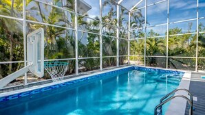 An indoor pool with clear glass walls, a basketball hoop, and a wooden deck. - 21431 Widgeon Bayfront Pool Home near the Beach (Fort Myers Beach)