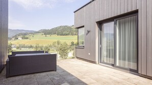 Ein Balkon mit einer Rattan-Couch, einer Glastür und Blick auf eine grüne Landschaft.