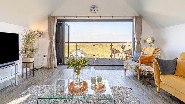A spacious living room with a large window, a flat-screen TV, a coffee table with decorative items, a sofa, and a dining area with chairs. - 3 Bed in Westward Ho! (oc-dreck) (Northam)