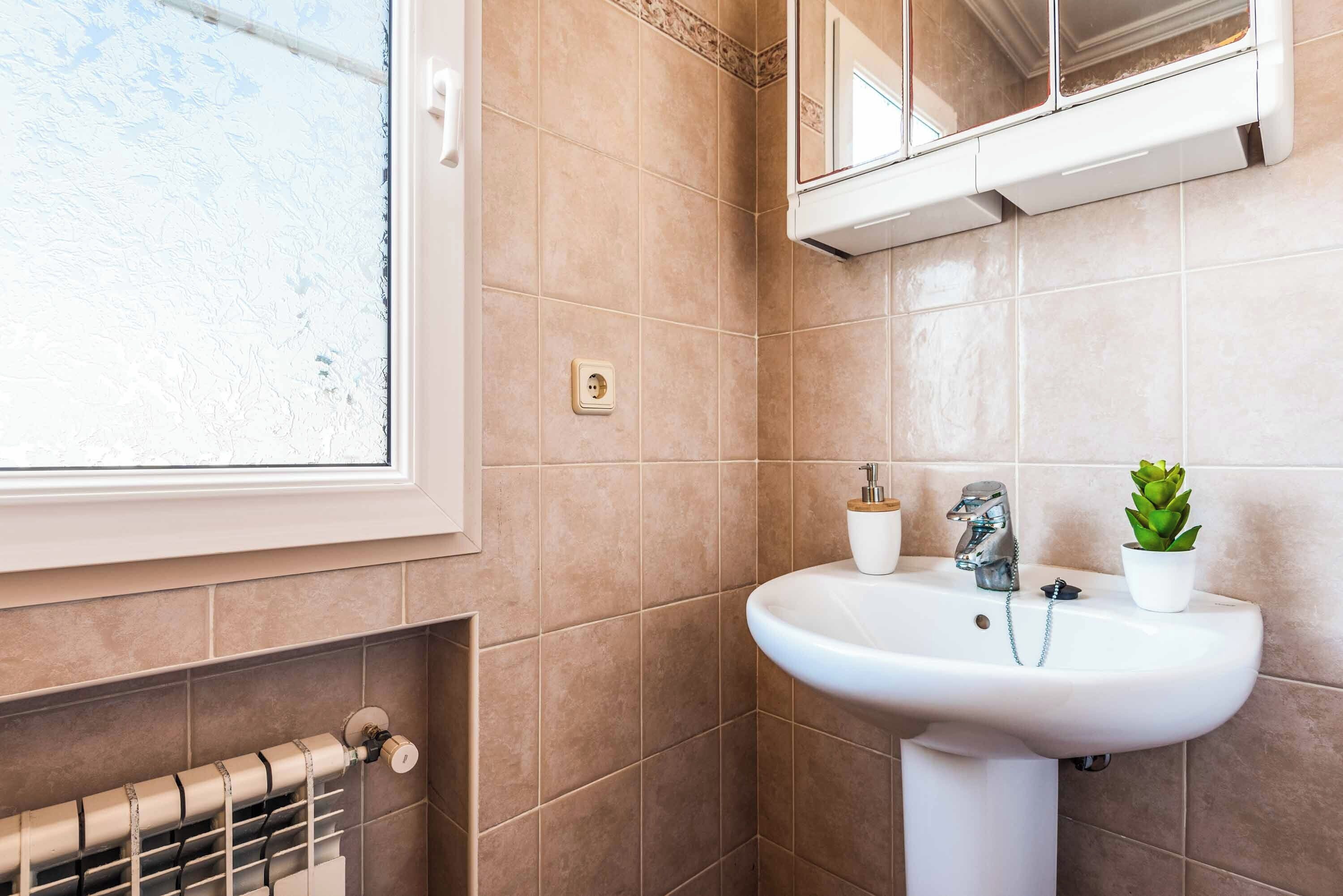 A bathroom with beige tiles, a white sink, a mirror, a window, and a radiator.