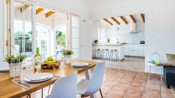 Un coin repas avec une table en bois dressée pour quatre personnes, des chaises blanches et, au fond, une cuisine avec des meubles blancs et un tabouret de bar.