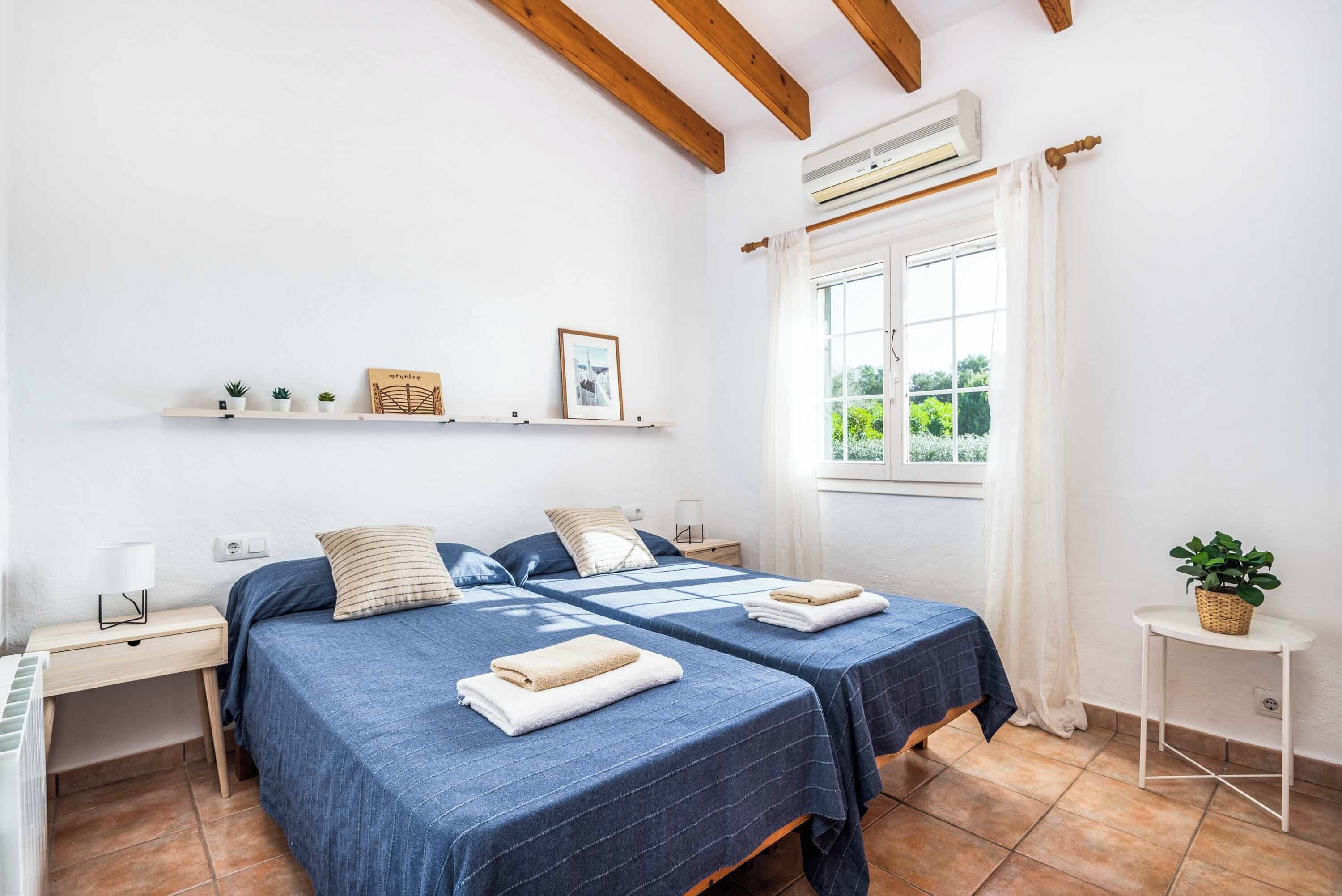 A room with two beds, a window with curtains, a small table with a plant, and a shelf with decorative items.