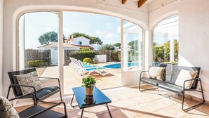 Une terrasse ensoleillée avec une piscine, deux chaises en métal noir, une petite table avec une plante, et une vue sur une maison blanche et l’espace piscine.