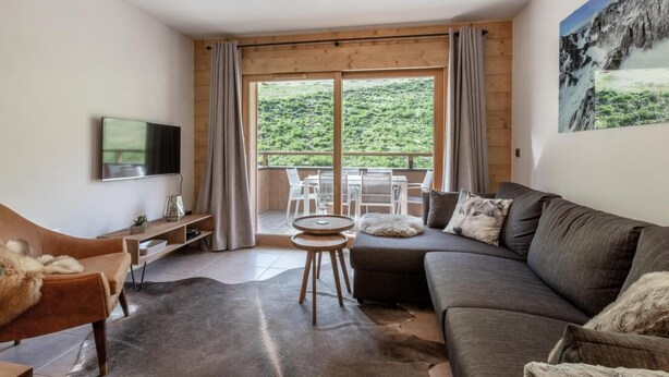 A modern living room with a large sofa, a TV, a small table, and a view of a green landscape through large windows.