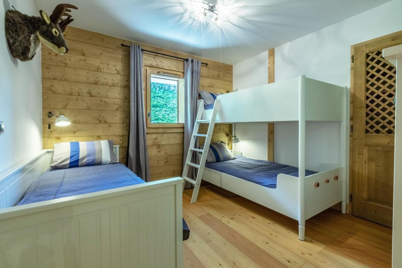 A room with two bunk beds, wooden walls, a window with curtains, and a mounted animal head.