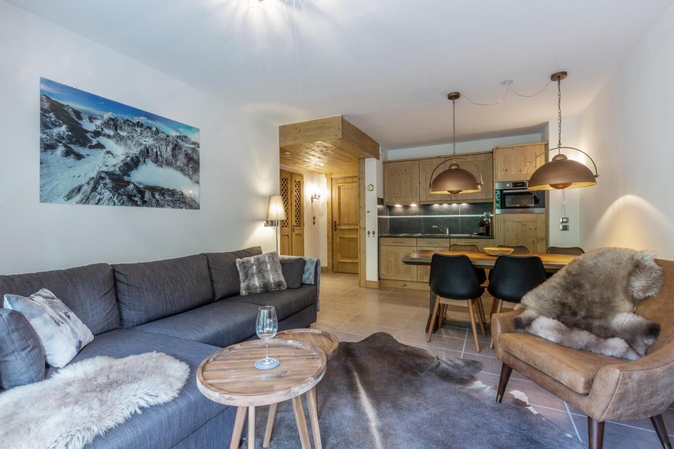 Ein modernes Wohnzimmer mit einer Couch, einem Couchtisch und einem Fellteppich. Im Hintergrund ist eine Küche mit Holdekor-Schränken und Pendelleuchten zu sehen.