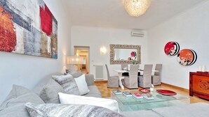 Ein modernes Wohnzimmer mit einem grauen Sofa, einem Glastisch und einem Essbereich mit weißem Tisch und grauen Stühlen.