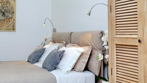 Ein ordentlich bezogenes Bett mit einer beige-weißen Daunendecke, eine Holztür mit horizontalen Lamellen und eine an der Wand befestigte Lampe.