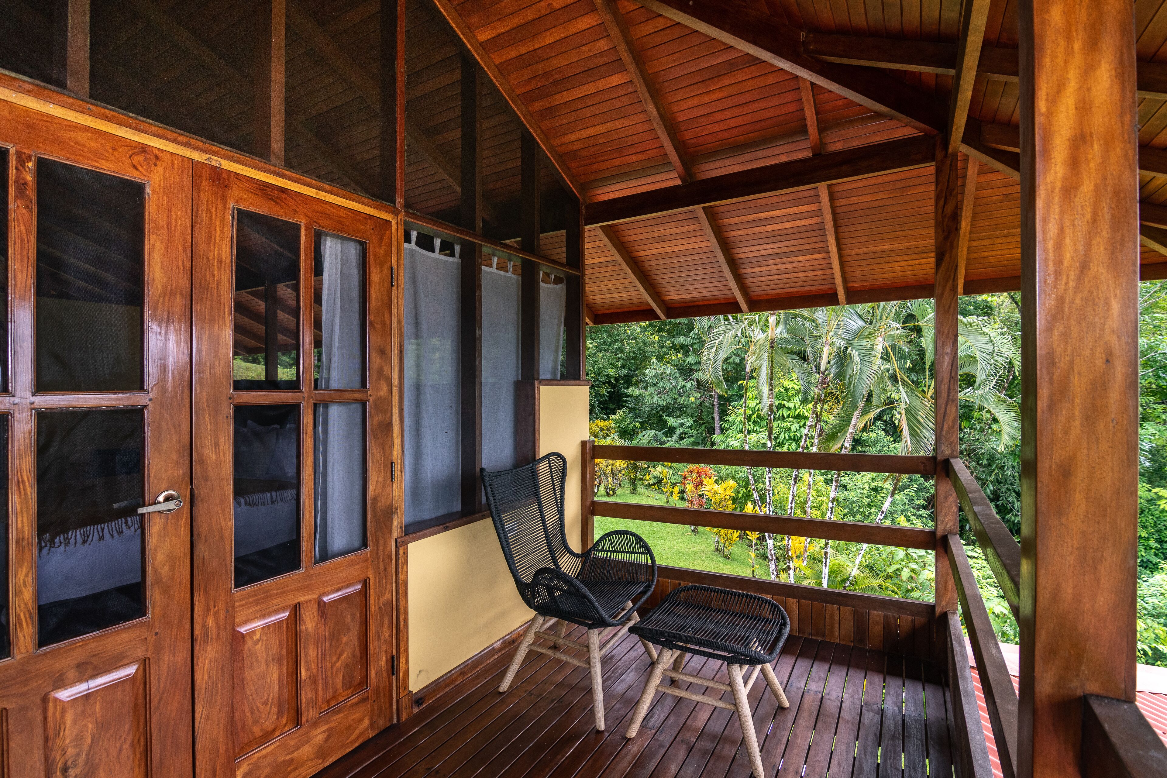 A wooden deck with a wooden ceiling, a black wicker chair, and a view of lush greenery.