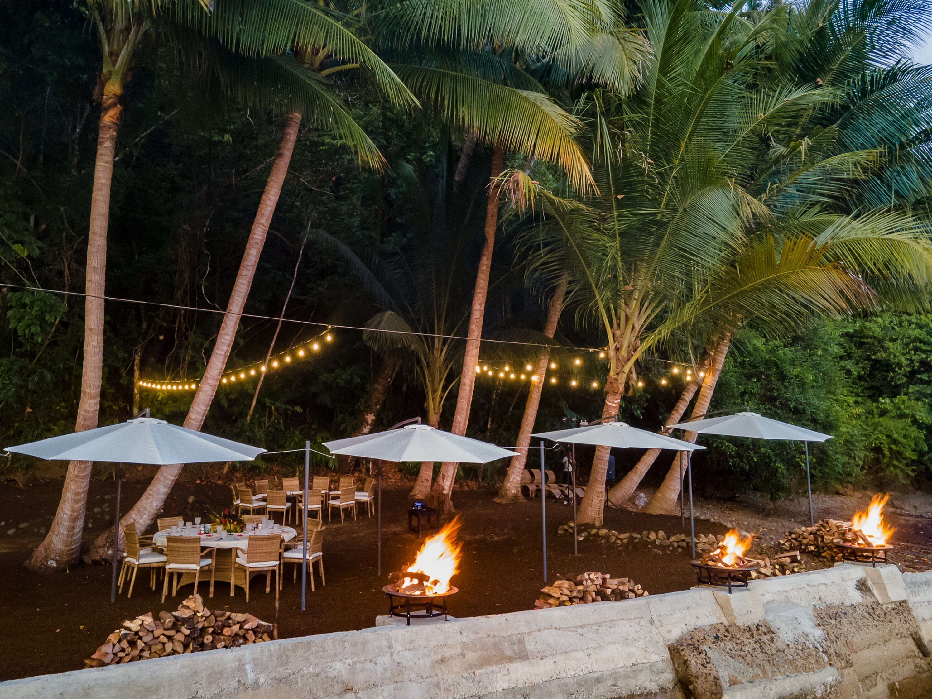 An outdoor dining area with tables and chairs, surrounded by palm trees and string lights.