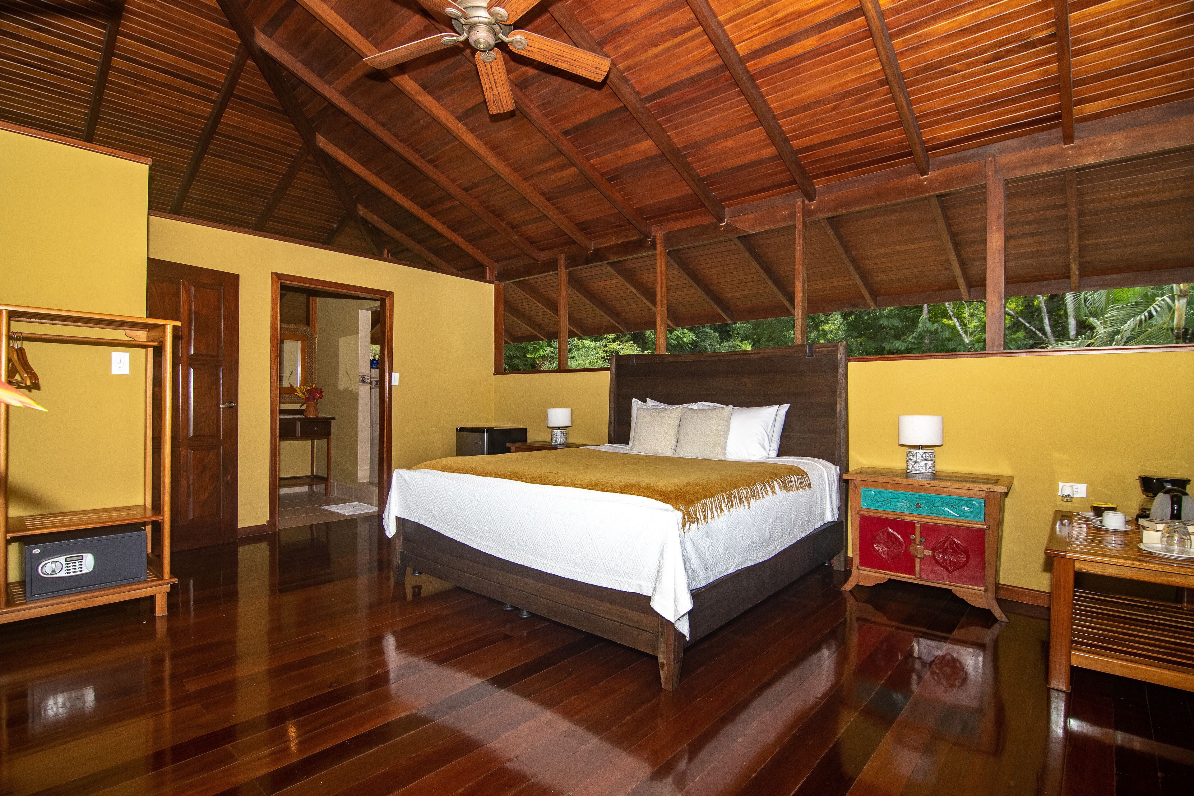 A spacious bedroom with a large bed, wooden ceiling, and a view of greenery outside.