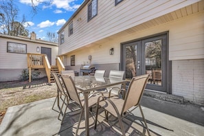 An outdoor patio with a glass-top table and metal chairs, a barbecue grill, and a wooden deck with a staircase.