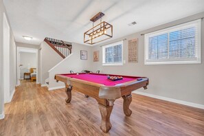 A pool table in a room with hardwood floors, a staircase, and a window with blinds.